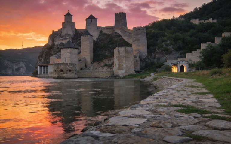 Golubačka tvrđava na obali Dunava pri zalasku sunca, kamene kule iznad reke i ulaz u Đerdapsku klisuru u pozadini.
